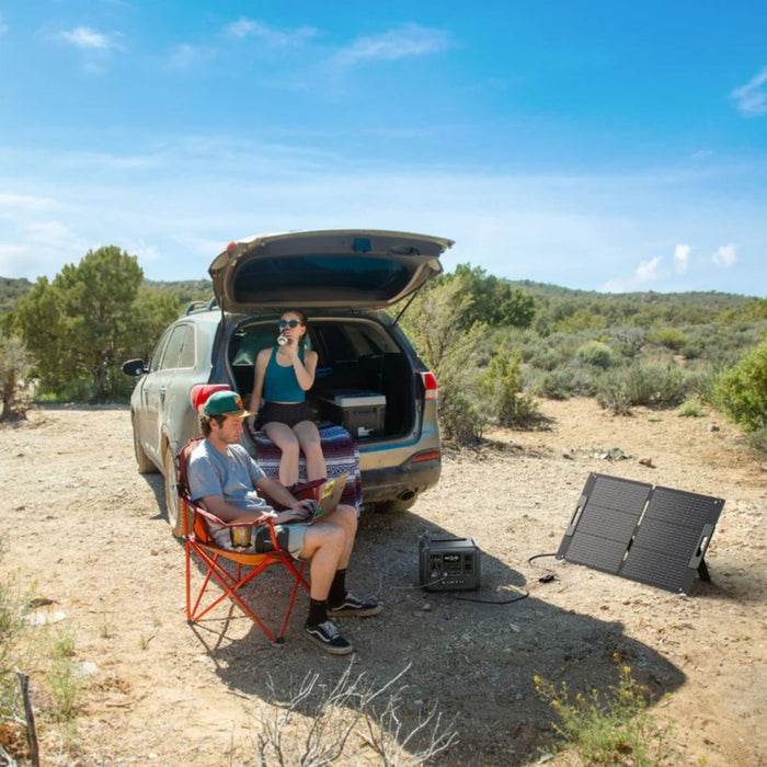 Two people in a desert setting with an open car trunk, Renogy Solar Blanket 400W 200W Portable Solar Panel, and camping chair.